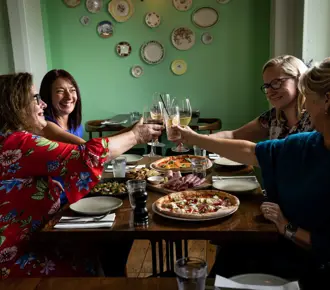 Group sit around a wooden table in Alpino restaurant in Cambridge, toasting with glasses of wine after exploring walking trails. The table overflows with pizza and appetizers. 