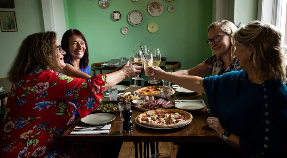 Group sit around a wooden table in Alpino restaurant in Cambridge, toasting with glasses of wine after exploring walking trails. The table overflows with pizza and appetizers. 