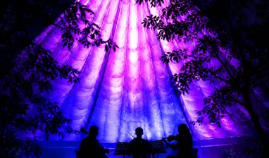 Three musicians perform on stage inside a large, glowing purple conical TreeChurch tent during an immersive light experience at night.