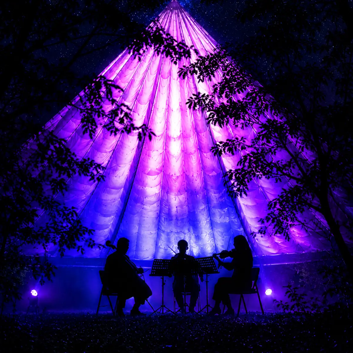 Three musicians perform on stage inside a large, glowing purple conical TreeChurch tent during an immersive light experience at night.