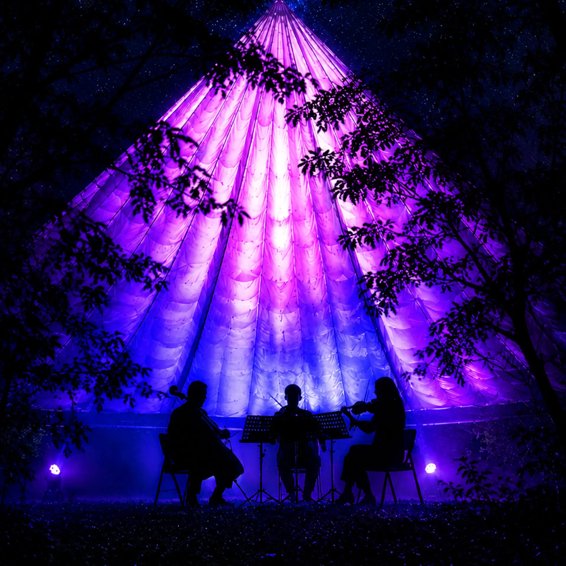 Three musicians perform on stage inside a large, glowing purple conical TreeChurch tent during an immersive light experience at night.