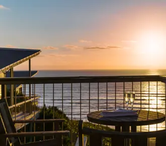 Two wooden chairs and a small round table on a balcony overlook the ocean at sunset at Castaway Resort. Two glasses are on the table—perfect for a couple's relaxing getaway. The sun casts a warm glow over the water and sky.