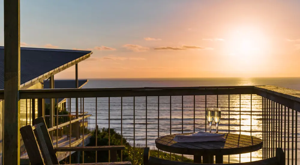 Two wooden chairs and a small round table on a balcony overlook the ocean at sunset at Castaway Resort. Two glasses are on the table—perfect for a couple's relaxing getaway. The sun casts a warm glow over the water and sky.