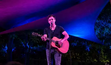 Man sings into a microphone while playing an acoustic guitar on an outdoor stage at the TreeChurch and Gardens, illuminated by an immersive light experience of blue and purple.